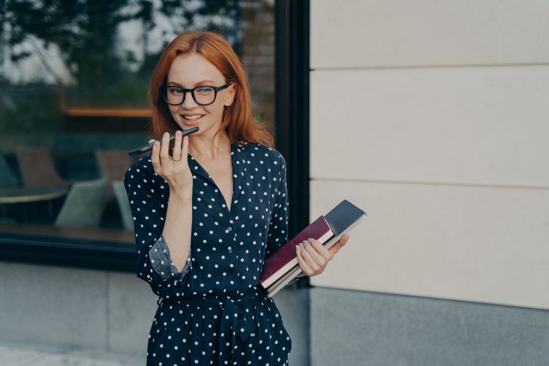 Professional woman using voice assistant while holding notebooks—representing efficient virtual assistant services for business support.